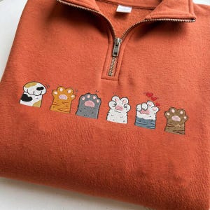 Könnte beinhalten: Ein rostorangefarbenes Sweatshirt mit Viertelreißverschluss und einer horizontalen Reihe aus sechs gestickten Katzenpfoten in verschiedenen Farben und Mustern, darunter Glückskatze, getigert, grau, weiß und braun, mit kleinen roten Herzen.