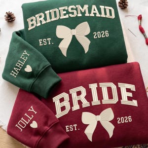 May include: A dark green sweatshirt with the text BRIDESMAID EST. 2026 and a white bow, and a maroon sweatshirt with the text BRIDE EST. 2026 and a white bow. The green sleeve features the name HARLEY with a heart, and the maroon sleeve says JOLLY with a heart.