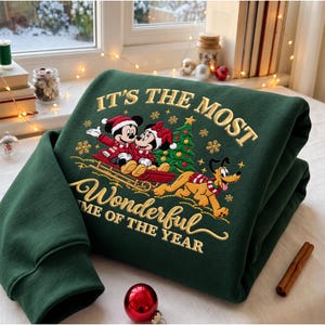 May include: A dark green sweatshirt featuring an embroidered graphic of Mickey and Minnie Mouse on a sled with Pluto running alongside. The design includes a Christmas tree, snowflakes, and the text IT'S THE MOST WONDERFUL TIME OF THE YEAR.