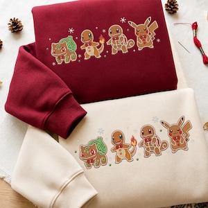 Può includere: Una felpa rosso scuro e una felpa color crema, entrambe caratterizzate da una stampa grafica di quattro personaggi in stile pan di zenzero che ricordano Bulbasaur, Charmander, Squirtle e Pikachu, decorati con piccoli fiocchi rossi e fiocchi di neve.