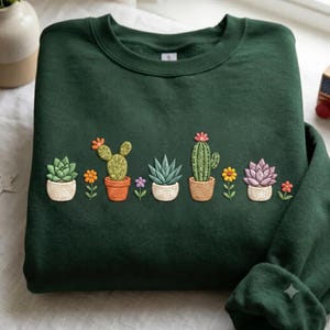 Op de afbeelding: Een donker bosgroene sweater met ronde hals voorzien van een borduursel van diverse cactussen en vetplanten in potten in tinten groen, oranje, paars en geel, gerangschikt in een horizontale rij over het midden van het kledingstuk.