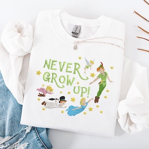 Op de afbeelding: Witte crewneck sweatshirt met de tekst "NEVER GROW UP!" in groen, met cartoon illustraties van Peter Pan, Tinkerbell, Wendy, John, Michael en Kapitein Haak. Een zilveren ketting met een hanger is ook zichtbaar.