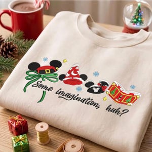 Könnte beinhalten: Ein beigefarbenes Sweatshirt mit Rundhalsausschnitt und einer Stickerei von Mickey-Maus-Ohren mit Weihnachtsmütze, einem Zaubererhut und einem Buch. Der Text "Some imagination, huh?" ist in schwarzer Schreibschrift unter den Motiven gestickt.