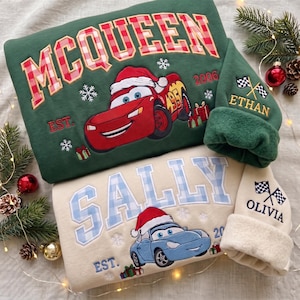 Op de afbeelding: Een donkergroen sweatshirt met een rode raceauto met kerstmuts en de tekst MCQUEEN EST. 2006, en een crèmekleurig sweatshirt met een blauwe auto met kerstmuts en de tekst SALLY EST. 2020. Beide mouwen tonen geblokte vlaggen en namen.
