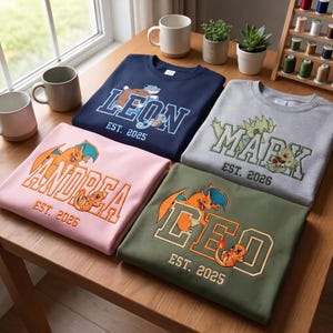 Peut inclure: Quatre sweat-shirts à col rond pliés en bleu marine, gris clair, rose clair et vert olive. Chacun présente des lettres brodées de style universitaire avec les noms LEON, MARK, ANDREA et LEO, accompagnés de graphismes de personnages et de dates EST.