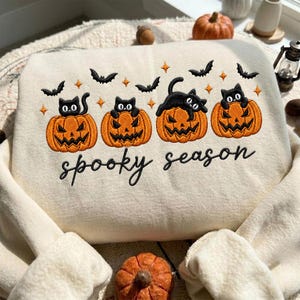Puede incluir: Una sudadera de color crema con un diseño bordado de cuatro calabazas de Halloween naranjas, cada una con un gato negro encima. El diseño incluye pequeños murciélagos negros y estrellas naranjas, con el texto "spooky season" bordado en negro.