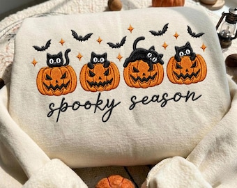 May include: A cream-colored sweatshirt featuring an embroidered design of four orange jack-o'-lanterns, each topped with a black cat. The design includes small black bats and orange stars, with the black embroidered text "spooky season" below.