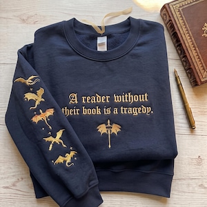 May include: A navy blue crewneck sweatshirt featuring gold embroidery. The chest displays the text "A reader without their book is a tragedy." with a dragon emblem below. The sleeve features a vertical sequence of five embroidered gold dragons.