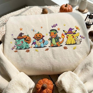 Peut inclure: Un sweat-shirt couleur crème avec un motif brodé représentant quatre créatures de dessin animé déguisées pour Halloween. Le design inclut une créature verte avec un chapeau de sorcière, une bleue avec une tête de citrouille, une verte en monstre et une jaune avec un chapeau de sorcière violet, entourées de bonbons et de feuilles.