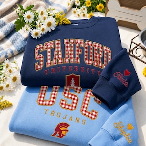 Könnte beinhalten: Ein marineblaues Sweatshirt mit STANFORD UNIVERSITY in rot-weiß karierten Buchstaben und einem Baum-Emblem sowie ein hellblaues Sweatshirt mit USC TROJANS in rot-weiß karierten Buchstaben und einem Trojaner-Helm-Emblem, beide mit Stickereien.
