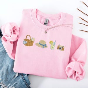 Op de afbeelding: Een lichtroze sweatshirt met geborduurde zomerse illustraties. Het ontwerp omvat een picknickmand, een zonnehoed met een blauw lint, een glas limonade en een vintage camera. Een zilveren ketting met een zonnehanger is ook zichtbaar.