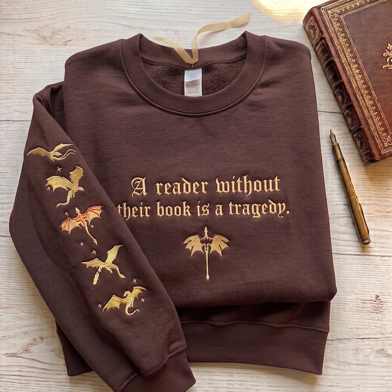 May include: A dark brown crewneck sweatshirt featuring gold embroidery. The chest displays the text "A reader without their book is a tragedy." with a sword icon below. The sleeve features five embroidered dragons in various flight poses.
