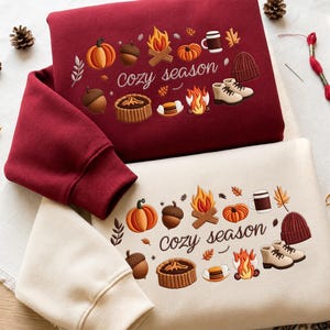 Może przedstawiać: Ciemnoczerwona bluza i kremowa bluza, każda z nadrukiem graficznym z napisem "cozy season" w otoczeniu ilustracji dyń, żołędzi, ognisk, kubka, czapki z dzianiny i butów w ciepłych jesiennych kolorach.