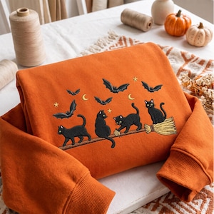 Op de afbeelding: Een oranje sweatshirt met een geborduurd ontwerp van drie zwarte katten en verschillende vleermuizen. Eén kat zit op een bezemsteel, terwijl andere eroverheen lopen. Het tafereel bevat kleine gele maansikkels en sterren op de stof.