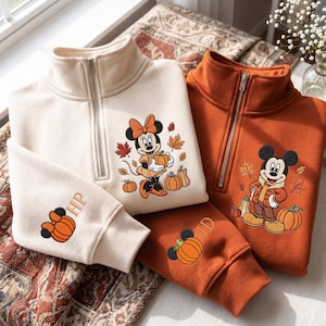 Puede incluir: Dos sudaderas con cremallera de cuarto, una color crema y otra naranja quemado, con personajes de Disney bordados. La sudadera crema muestra a Minnie Mouse con calabazas y hojas, mientras que la naranja presenta a Mickey Mouse. Ambas mangas tienen iconos de calabaza con las iniciales HP y JD.