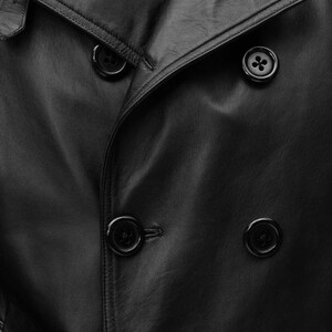 May include: A close-up shot of a black leather coat with a double-breasted design. The coat has large, round black buttons and a smooth, textured surface. The image focuses on the details of the coat's construction and material.