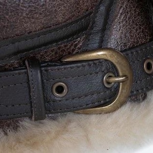 May include: Close-up of a brown leather aviator hat with a dark brown leather strap and a brass buckle. The hat has a textured appearance and is lined with a cream-coloured, fluffy material.
