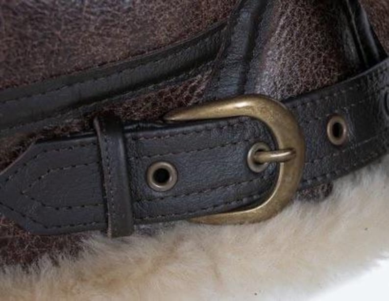 May include: Close-up of a brown leather aviator hat with a dark brown leather strap and a brass buckle. The hat has a textured appearance and is lined with a cream-coloured, fluffy material.