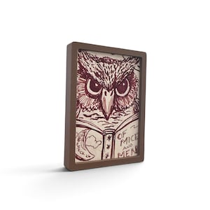 May include: A rectangular brown frame containing a drawing of an owl reading a book. The illustration is rendered in dark red ink on a light background. The book cover features the text Of Mice and Men and a crescent moon with the text ESECK 26.
