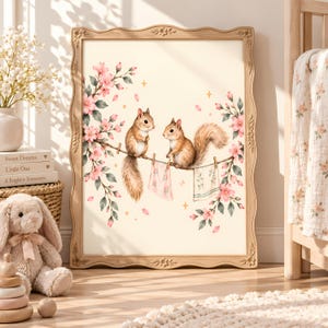 Könnte beinhalten: Ein gerahmter Kunstdruck mit zwei braunen Eichhörnchen auf einem Ast, umgeben von rosa Kirschblüten und zwei kleinen Stoffstücken, die an Wäscheklammern hängen. Der Hintergrund ist cremefarben und mit kleinen goldenen Sternen verziert.