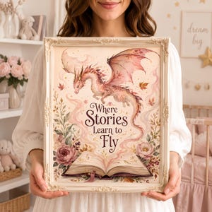 May include: A framed wall art print featuring a watercolour-style illustration of a pink dragon flying above an open book. The text "Where Stories Learn to Fly" is centred in dark brown script. The artwork includes floral accents and autumn leaves.