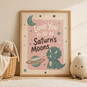 Könnte beinhalten: Ein gerahmter Wandkunstdruck mit einem rosafarbenen, gewellten Rechteck und dem Text Love You To All Of Saturn's Moons. Das Design zeigt eine türkisfarbene Mondsichel, einen gestreiften Planeten mit Ringen und einen lächelnden grünen Triceratops.