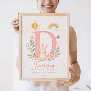 Puede incluir: Una impresión en acuarela enmarcada con la letra "D" rosa, un conejito, flores, un arcoíris y un sol. El nombre "Donna" está escrito debajo, con el texto "Dream Big, Little One" y "You are so loved".