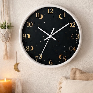 Könnte beinhalten: Eine runde Wanduhr mit weißem Rahmen. Das schwarze Zifferblatt hat goldfarbene Mondphasenzahlen und Sternakzente. Weiße Zeiger zeigen die Uhrzeit an. Die Uhr ist ein dekorativer Artikel, ca. 30 cm im Durchmesser.