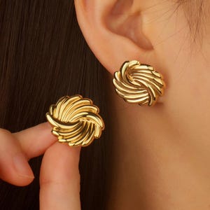 May include: A pair of gold-coloured stud earrings. Each earring has a swirling, textured design. One earring is being held by a person's fingers, while the other is on the ear. The earrings have a shiny finish.