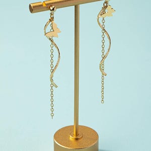 May include: Gold-colored earrings with a butterfly design. Each earring features a butterfly charm, a curved, wavy bar, and a delicate chain. The earrings are displayed on a gold-colored stand against a light blue background.