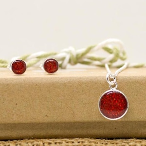 May include: A jewellery set comprising a necklace and stud earrings. The pendant and earrings feature a round, silver setting filled with sparkling red glitter. The necklace has a delicate silver chain.