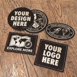 May include: Five black patches with white engraved designs. One circular patch reads YOUR DESIGN HERE, another features a rocket and parachute. An oval patch reads YOUR ADVENTURE with coordinates. A rectangular patch shows a map and compass with EXPLORE MORE, and a square patch reads YOUR LOGO HERE.
