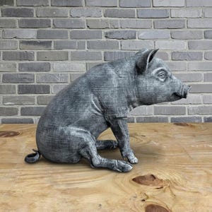 May include: A gray-toned pig sculpture in a seated position, with its front legs extended and its tail curled. The pig has detailed features and is set against a wooden surface and a gray brick wall.