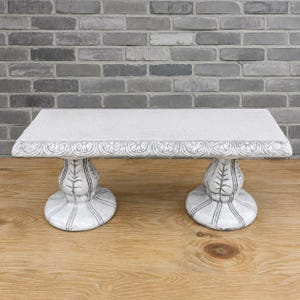 May include: A rectangular stone display stand with a textured, light gray surface and a decorative scalloped edge. The stand is supported by two matching pedestal bases, each featuring a carved, ribbed design with a wide, circular foot.