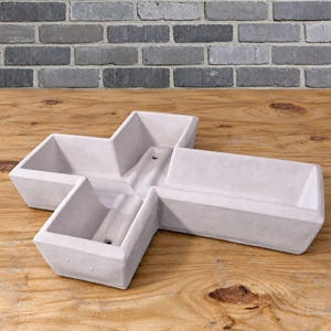 May include: A light gray concrete planter shaped like a cross with four distinct rectangular compartments. The structure features a smooth, matte finish and includes small drainage holes at the base of each section, creating a geometric vessel.