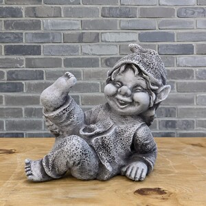 May include: A gray stone garden gnome figurine depicted in a seated position with one leg raised high. The gnome features a textured pointed hat, a wide smiling face with prominent cheeks, and detailed clothing with a stippled surface finish.