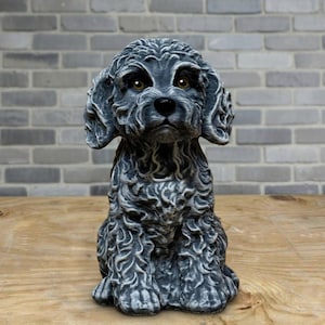 May include: A detailed, gray dog statue with a curly coat and attentive expression. The dog is seated, with its front paws visible. The statue has brown eyes and is set against a brick wall and wooden surface.