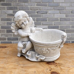 May include: A light gray stone planter featuring a sculpted cherub figure with wings leaning against a round basin. The basin is decorated with a relief of draped fabric and floral garlands, showcasing a weathered texture and a matte finish.