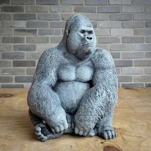 May include: A gray gorilla statue in a seated position, with a serious expression. The statue has detailed fur texture and is displayed on a wooden surface against a brick wall background. The gorilla's chest and arms are prominently featured.