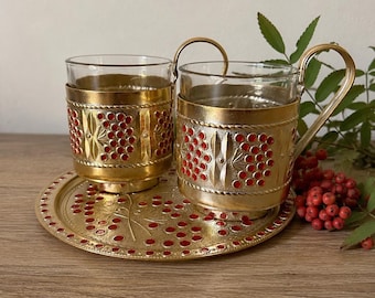 May include: Two glass teacups with gold-coloured metal holders and handles, on a matching tray. The holders and tray feature a red berry design. The set is displayed on a wooden surface.