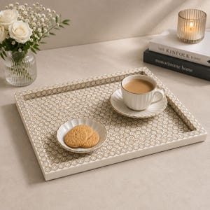 May include: A rectangular serving tray with a white frame and a repeating beige and white geometric hexagon pattern on the base. The tray holds a white ceramic teacup on a matching saucer and a small scalloped dish containing two round cookies.
