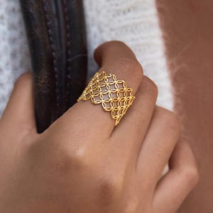 May include: A wide gold ring featuring an intricate, openwork lattice pattern with a scalloped edge. The band is constructed from a repeating geometric grid of small, textured circular links, creating a delicate, lace-like appearance on the finger.