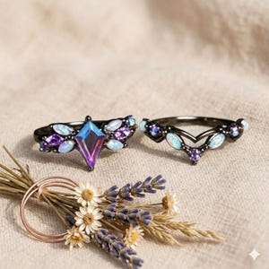 May include: Two black metal rings featuring iridescent kite-shaped and marquise-cut stones in shades of purple and blue, accented with small white opalescent gems. The rings are displayed alongside a small bundle of dried lavender and daisies.