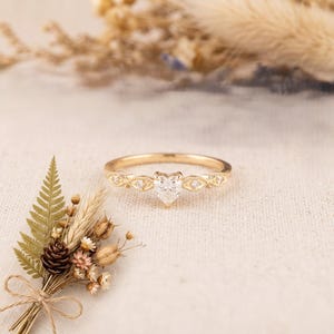 Pode incluir: Um anel de ouro com uma pedra preciosa transparente em forma de coração no centro, ladeada por duas pedras redondas mais pequenas de cada lado, montado numa banda delicada com detalhes milgrain ao longo das bordas metálicas dos engastes.