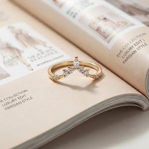 Peut inclure: Une bague en or en forme de V ornée d'une pierre centrale taille marquise, entourée de quatre petites pierres rondes de chaque côté. L'anneau est en or poli et les pierres sont transparentes et facettées, formant un motif symétrique.