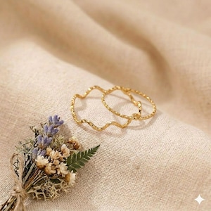 May include: Two thin gold rings with a wavy, textured surface. One ring features a continuous undulating design, while the other is a simple circular band with a hammered finish. A small bouquet of dried lavender and white flowers is nearby.