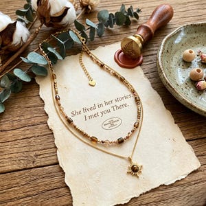 May include: A layered gold necklace with brown and clear beads and a sunburst pendant. The necklace rests on a piece of paper with the text "She lived in her stories. I met you There."