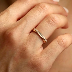 Könnte beinhalten: Ein Goldring bestehend aus zwei miteinander verbundenen Bändern. Ein Band hat eine glatte, polierte Goldoberfläche, während das andere Band schmaler ist und mit einer Reihe kleiner, klarer, funkelnder Steine auf der Oberseite besetzt ist.