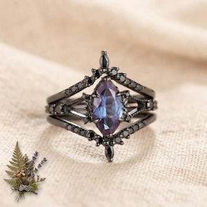 Könnte beinhalten: Ein Ring aus schwarzem Metall mit einem zentralen, marquise-geschliffenen Stein, der zwischen Violett und Blau changiert. Die Ringschiene ist mit kleinen, runden schwarzen Steinen besetzt, die in den filigranen, spitz zulaufenden Rahmen eingefasst sind.