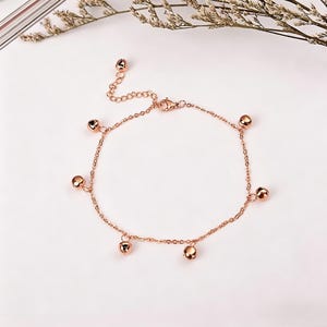 May include: Rose gold anklet featuring a delicate chain design. The anklet is adorned with several small, round, rose gold-colored charms. The jewelry is displayed on a white surface, showcasing its elegant simplicity.
